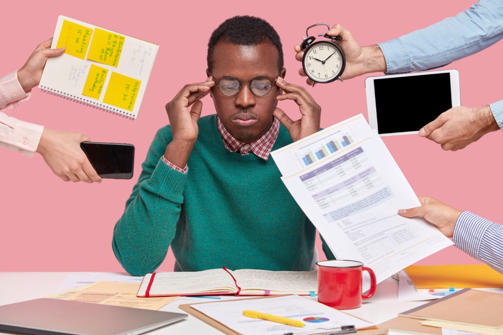 A stressed man sitting at a desk, surrounded by multiple hands offering documents with management accounts, gadgets, and a clock, illustrating the overwhelm of multitasking and managing various tasks simultaneously.