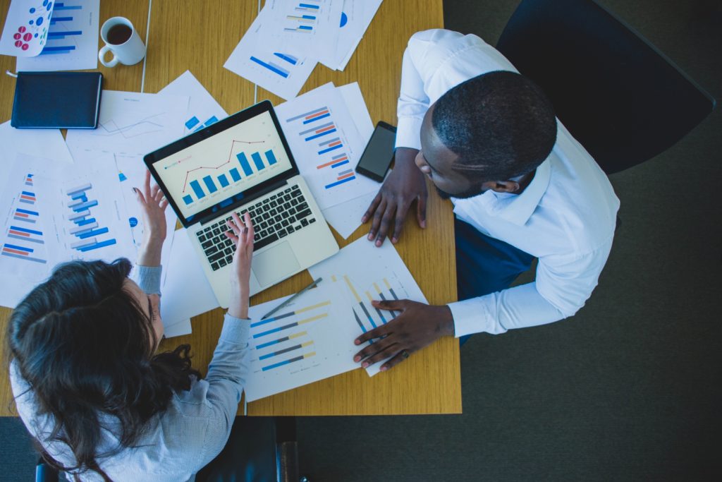 Two business professionals reviewing financial charts and graphs on a laptop and printed documents, analyzing data to make informed decisions.